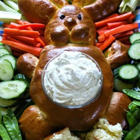 bread shaped like bunny, surrounded by raw cucumbers, bell peppers, broccoli, radishes, and carrots with their stems attached, belly of bread bunny lined with lettuce leaves and filled with dip