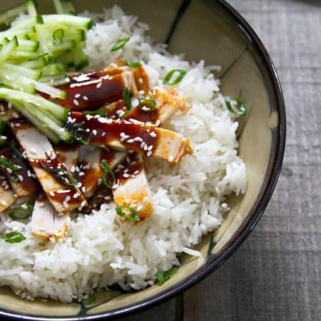 Strips of slow-cooker korean style barbecue pork with sauce on rice with cucumbers in taupe ceramic bowl with a black rim, on wood background.