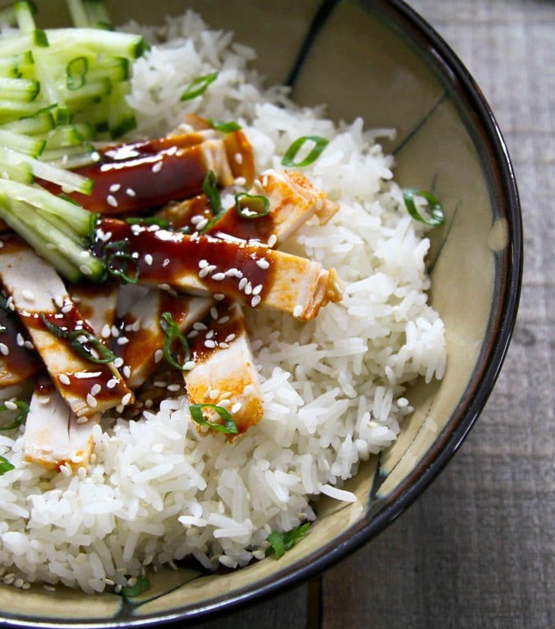 Strips of slow-cooker korean style barbecue pork with sauce on rice with cucumbers in taupe ceramic bowl with a black rim, on wood background.