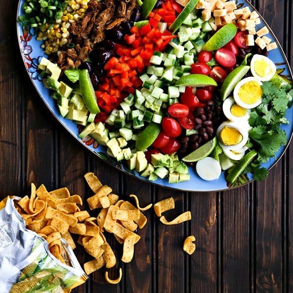 Square crop Tex-Mex Cobb Salad on blue floral Mackenzie Childds platter, open bag of fritos, dark wood table, greens, corn, cucumbers, black beans, hard boiled eggs, carnitas, cherry tomatoes, red bell peppers, blackk olives, cubed cheese, avocados.