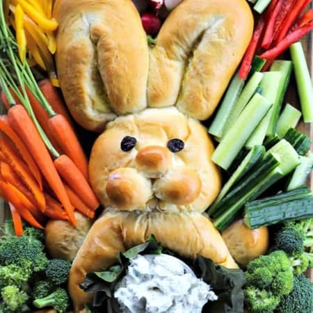 bread shaped like bunny, surrounded by raw cucumbers, bell peppers, broccoli, radishes, and carrots with their stems attached, belly of bread bunny lined with lettuce leaves and filled with dip