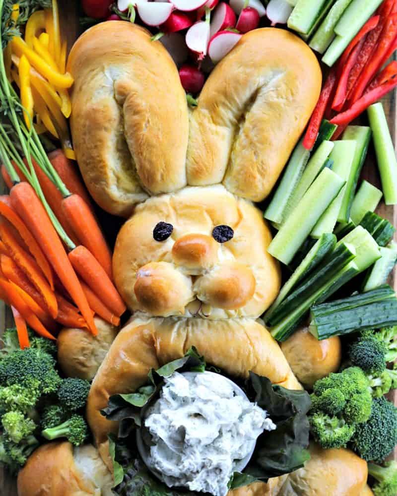 bread shaped like bunny, surrounded by raw cucumbers, bell peppers, broccoli, radishes, and carrots with their stems attached, belly of bread bunny lined with lettuce leaves and filled with dip