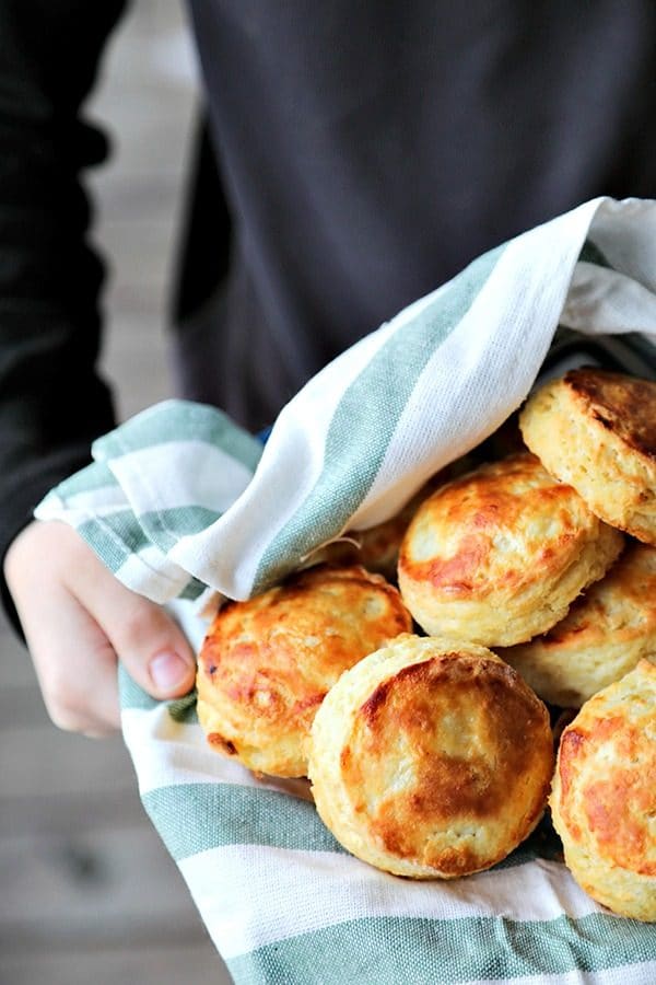 perfect flaky buttermilk biscuits in green and white towel lined bowl, held by boy in black long sleeved shirt, wooden porch