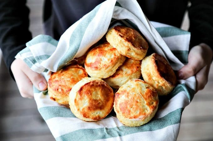 perfect flaky buttermilk biscuits in green and white towel lined bowl, held by boy in black long sleeved shirt, wooden porch