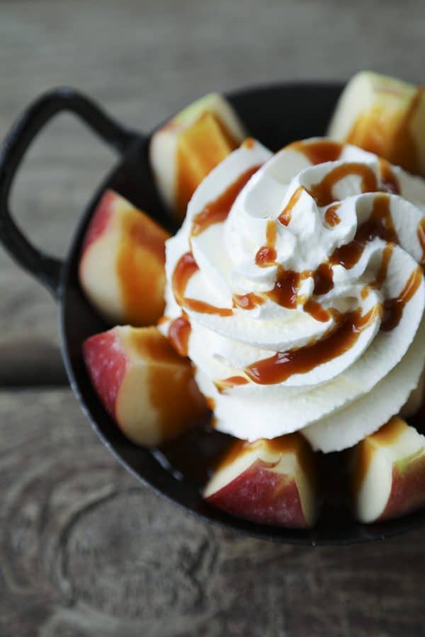 2-Minute Pull Apart Caramel Apples , apple wedges, whipped cream topped with dripping caramel sauce and cinnamon, black and white cast enamel bowl with handles, wooden table