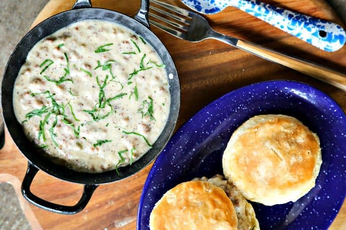 Classic Sausage Gravy in black and white enameled bowl with handles, biscuits on blue and white enamel plate, round wooden platter, fork with wooden handle, blue and white enamel metal spoon