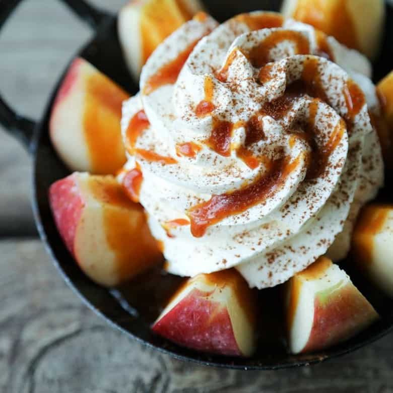 2-Minute Pull Apart Caramel Apples , apple wedges, whipped cream topped with dripping caramel sauce and cinnamon, black and white cast enamel bowl with handles, wooden table