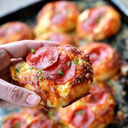 Pepperoni Pizza Bialys Recipe held by hand, melted cheese, crisp pepperoni, parsley, background of pizza on antique cookie sheet.