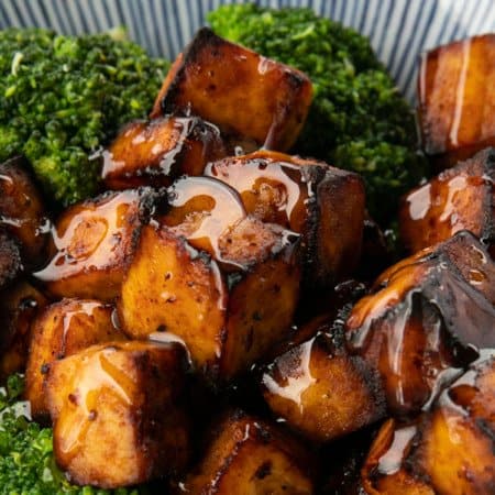 Crispy, golden brown cubes of air fryer tofu served with a drizzle of honey and broccoli on a blue and white striped oblong bowl.