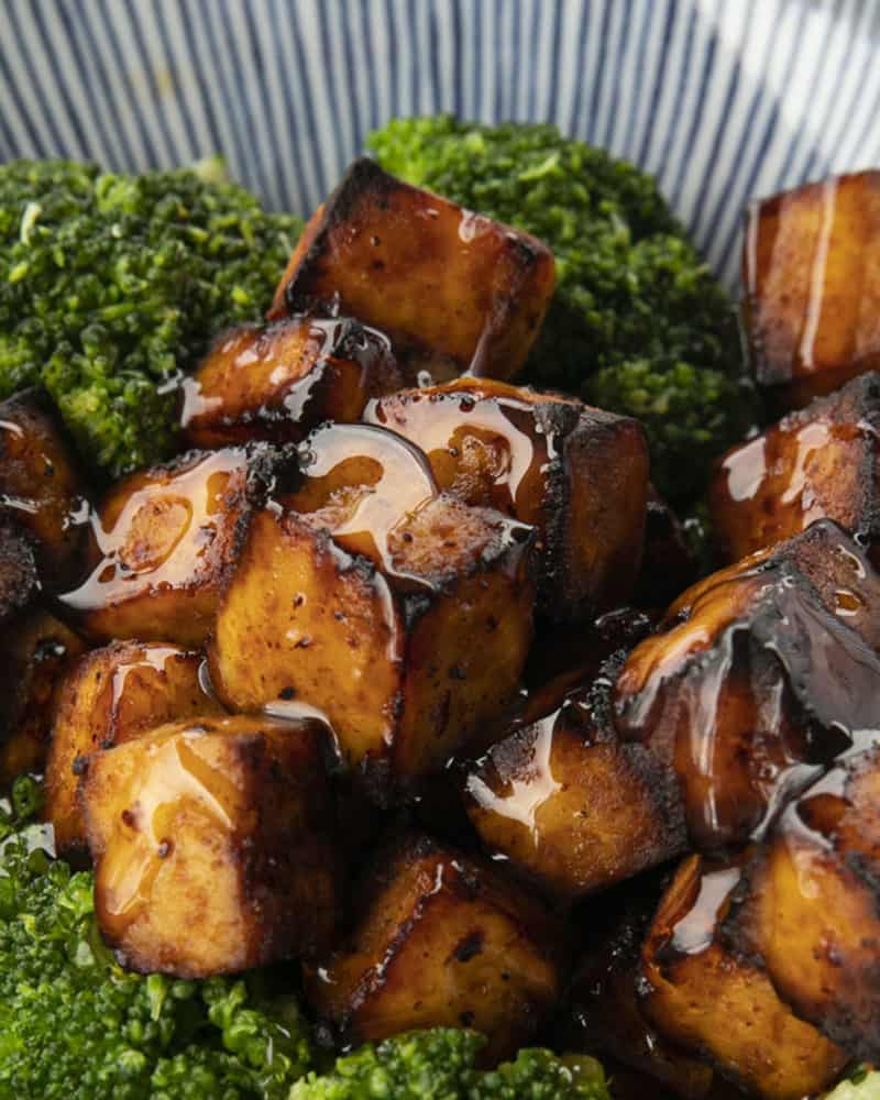 Crispy, golden brown cubes of air fryer tofu served with a drizzle of honey and broccoli on a blue and white striped oblong bowl.