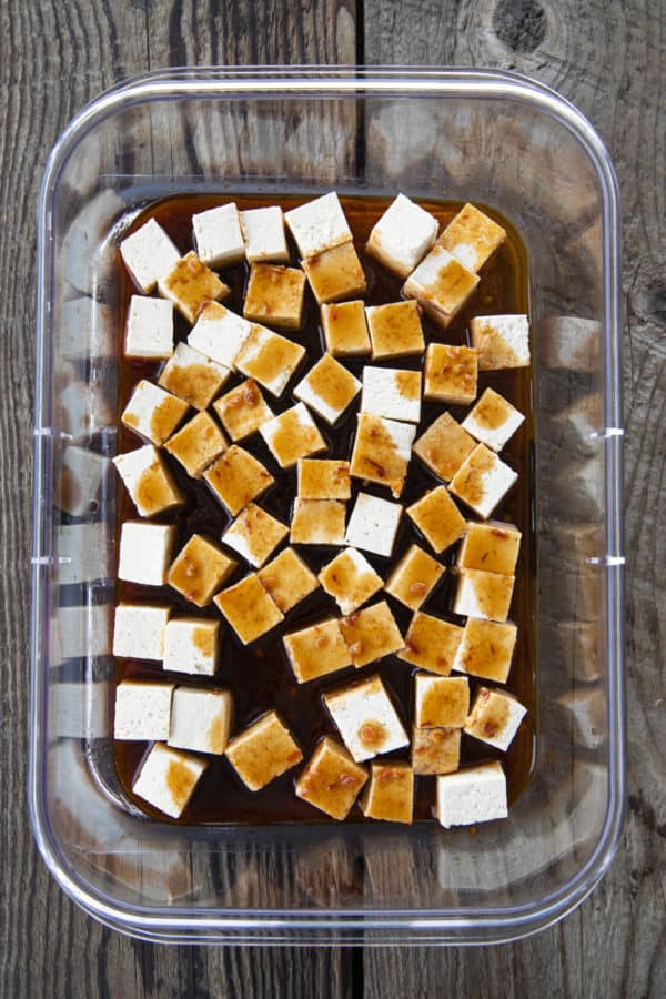 Small cubes of firm tofu in marinade in a transparent, rectangular container.