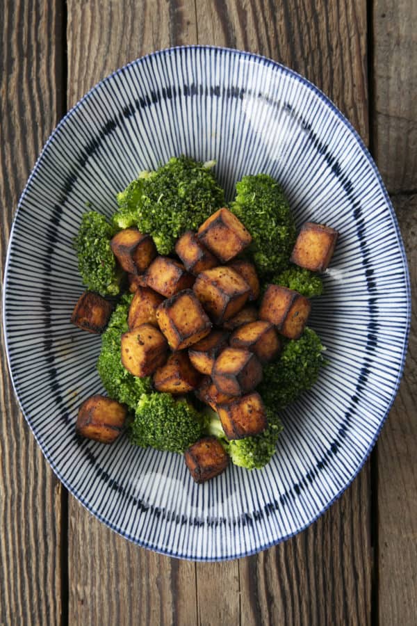 Crispy, golden brown cubes of air fryer tofu served with a drizzle of honey and broccoli on a blue and white striped oblong bowl.