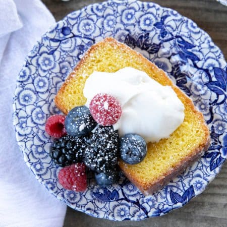French Grandma lemon yogurt cake with berries, powdered sugar, and yogurt on a blue and white plate with a white linen.
