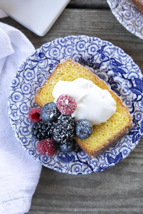 French Grandma lemon yogurt cake with berries, powdered sugar, and yogurt on a blue and white plate with a white linen.