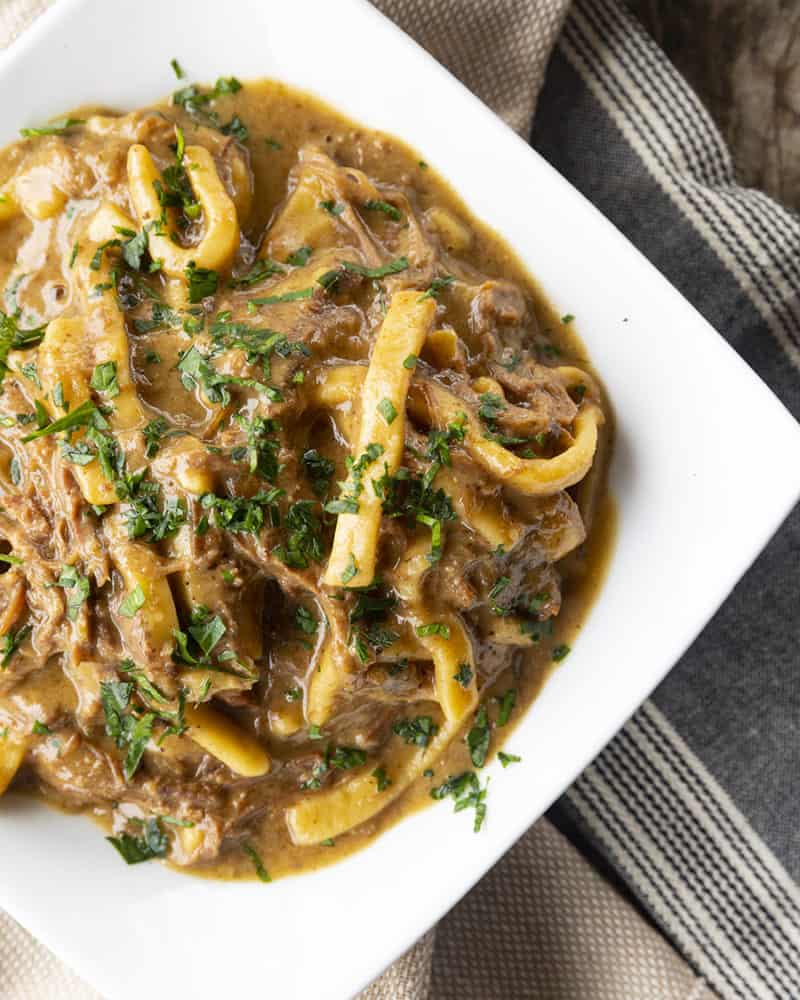 crock pot beef and noodles in a white square bowl on linens