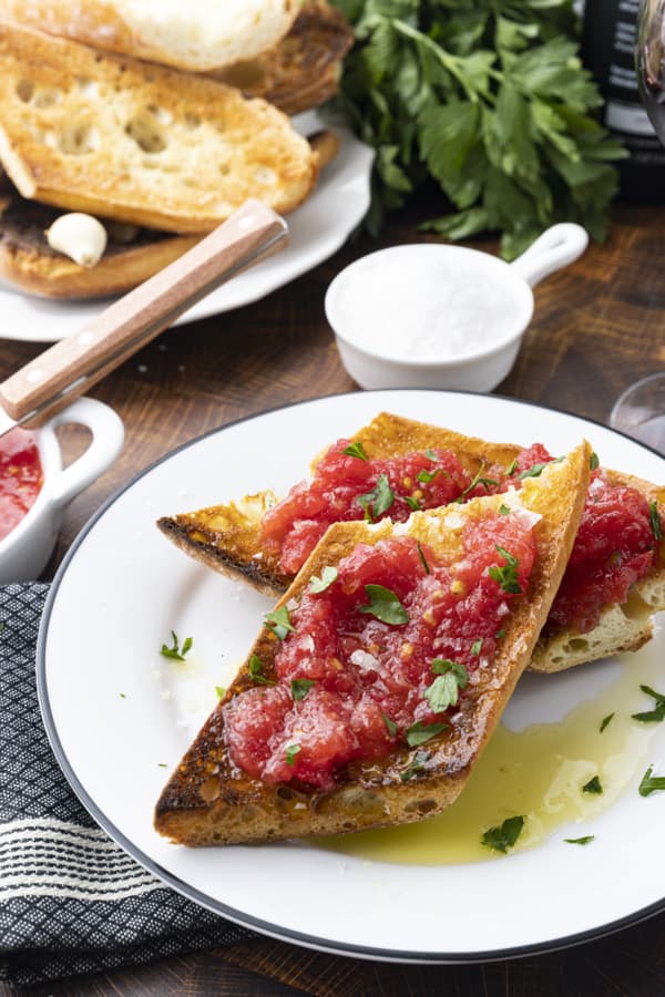 Sublime and simple Pan Con Tomate has just five ingredients; toasted rustic bread rubbed with a garlic clove, juicy fresh tomato pulp, extra-virgin olive oil, and coarse flaky sea salt. Also known as Spanish Tomato Bread, Pan Tumaca, or Pa Amb Tomàquet, this easy recipe using simple ingredients is equally at home served at a dinner party with a Spanish omelette or as part of a traditional Spanish breakfast.