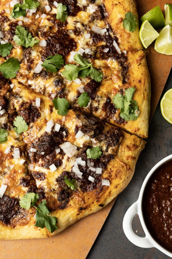 Birria Pizza: Chewy crust topped with tender shredded birria beef, smoky birria stew, loads of melted cheese, chopped sweet onion, fresh cilantro, a squeeze of fresh lime, and a bowl of red chile sauce for dipping. Imagine the best tacos you’ve ever tasted turned into a pizza and you have an idea just how fabulous this pizza is.