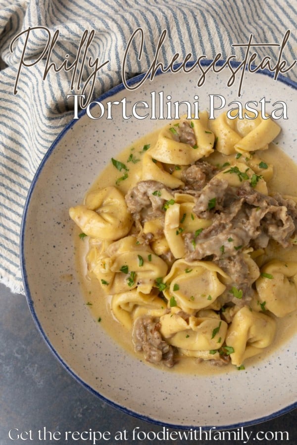 Philly cheesesteak tortellini in rich provolone sauce garnished with chopped parsley in a blue rimmed speckled bowl on a table with a blue and white striped linen with text overlay describing the photo