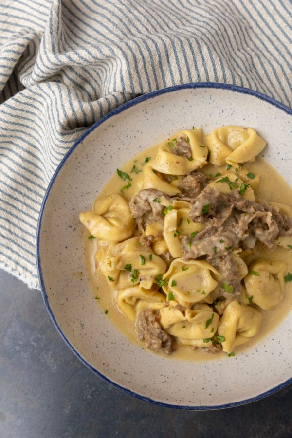 Philly cheesesteak tortellini in rich provolone sauce garnished with chopped parsley in a blue rimmed speckled bowl on a table with a blue and white striped linen