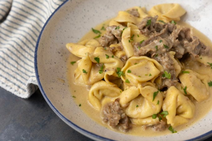 Philly cheesesteak tortellini in rich provolone sauce garnished with chopped parsley in a blue rimmed speckled bowl on a table with a blue and white striped linen
