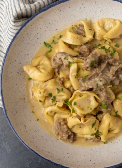 Philly cheesesteak tortellini in rich provolone sauce garnished with chopped parsley in a blue rimmed speckled bowl on a table with a blue and white striped linen