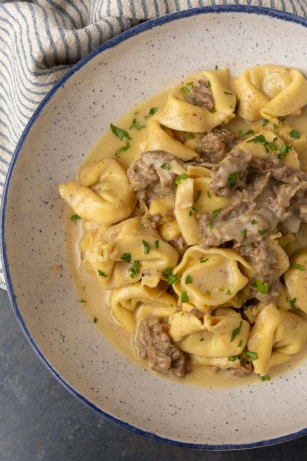 Philly cheesesteak tortellini in rich provolone sauce garnished with chopped parsley in a blue rimmed speckled bowl on a table with a blue and white striped linen
