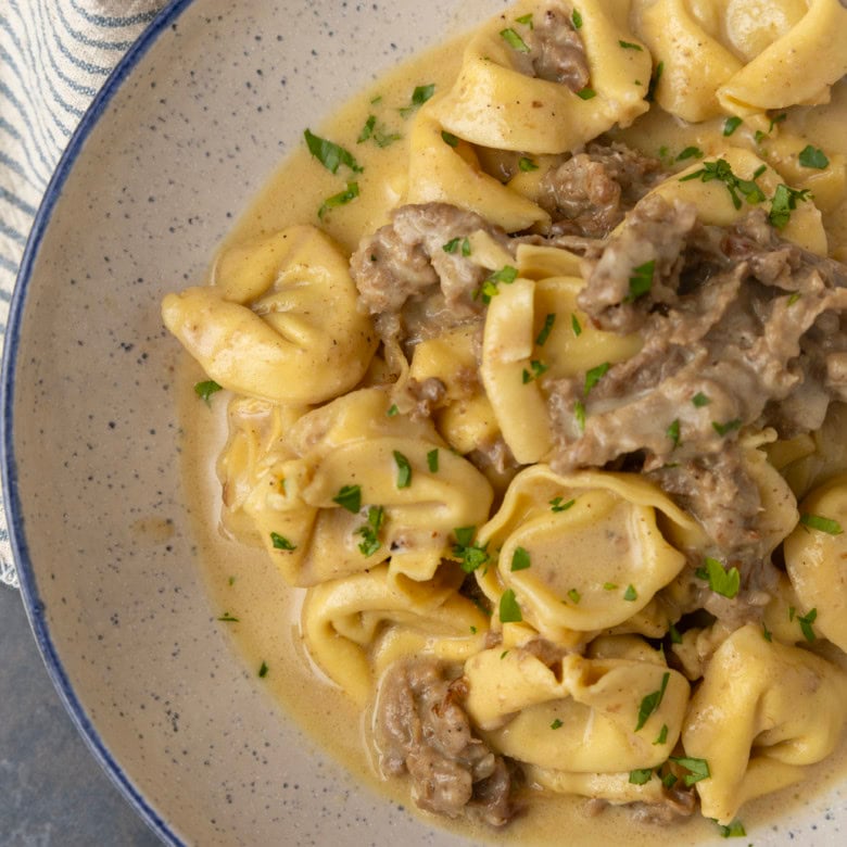 Philly cheesesteak tortellini in rich provolone sauce garnished with chopped parsley in a blue rimmed speckled bowl on a table with a blue and white striped linen