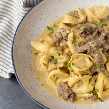 Philly cheesesteak tortellini in rich provolone sauce garnished with chopped parsley in a blue rimmed speckled bowl on a table with a blue and white striped linen