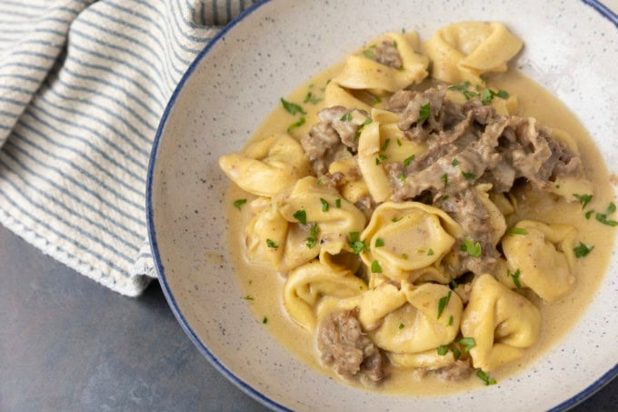 Philly cheesesteak tortellini in rich provolone sauce garnished with chopped parsley in a blue rimmed speckled bowl on a table with a blue and white striped linen