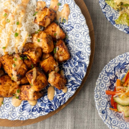 crispy air fryer bang bang salmon bites drizzled with sauce served with ice on a blue and white floral plate and two smaller blue and white plates with Japanese cabbage salad and pickled pepper salad