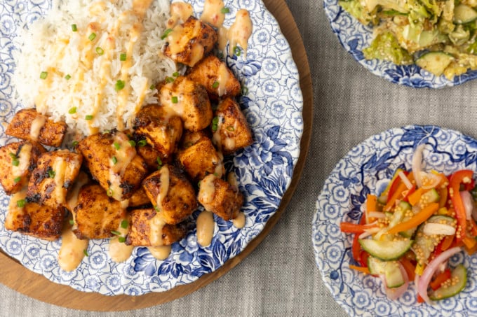crispy air fryer bang bang salmon bites drizzled with sauce served with ice on a blue and white floral plate and two smaller blue and white plates with Japanese cabbage salad and pickled pepper salad