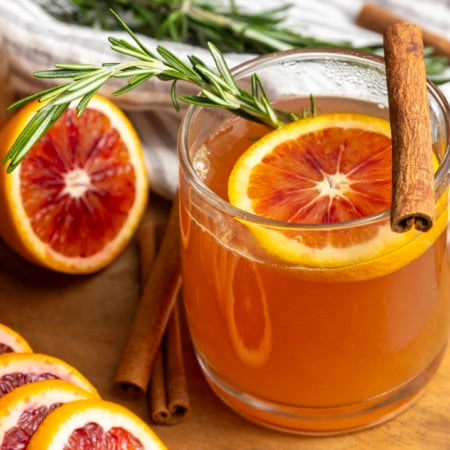 Orange tea in a mug with a slice of orange, a cinnamon stick, and a sprig of rosemary.