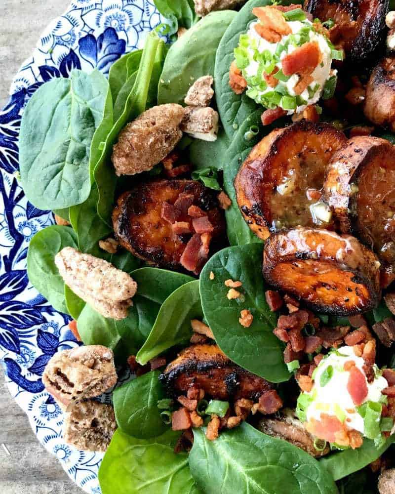 Melting Sweet Potato Salad, baby spinach, caramelized roasted sweet potatoes, maple candied pecans, crumbled bacon, goat cheese with herbs and bacon, chopped chives, on an antique blue and white plate.