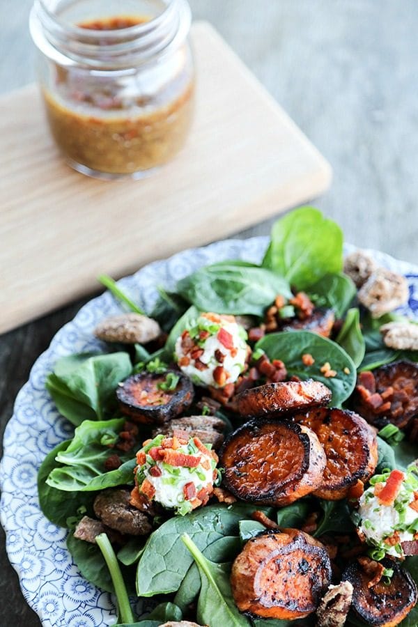 Melting Sweet Potato Salad, baby spinach, caramelized roasted sweet potatoes, maple candied pecans, crumbled bacon, goat cheese with herbs and bacon, chopped chives, on an antique blue and white plate, wooden cutting board with mason jar filled with hot bacon vinaigrette