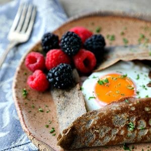 Buckwheat Crepe with fried egg, ham, and cheese on a pottery plate that looks like wood, fresh blackberries, parsley garnish