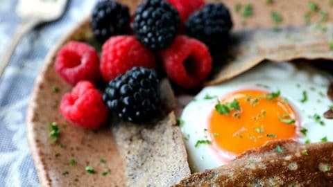 Buckwheat Crepe with fried egg, ham, and cheese on a pottery plate that looks like wood, fresh blackberries, parsley garnish