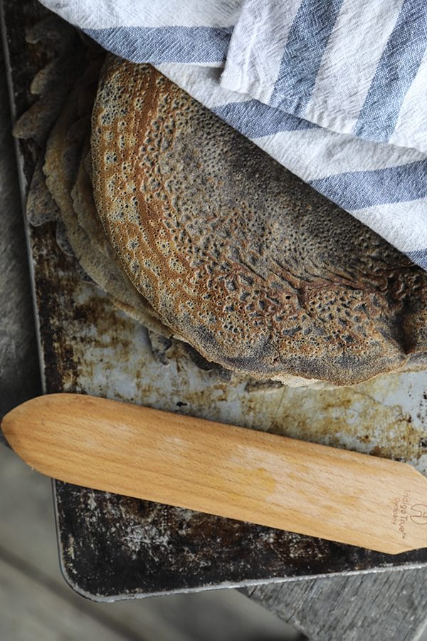 Pure buckwheat crepes on an antique cookie sheet under a blue and white towel, wooden crepe turner