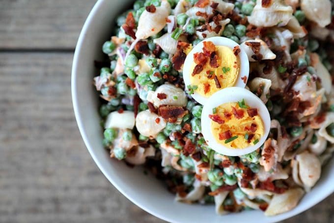 bacon pea pasta salad in white bowl with hard boiled eggs, bacon bits, rustic wood table