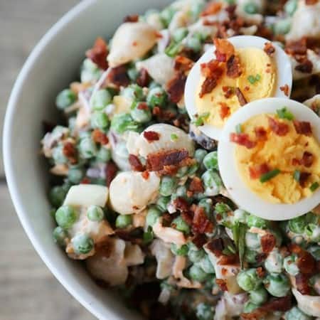 bacon pea pasta salad in white bowl with hard boiled eggs, bacon bits, rustic wood table