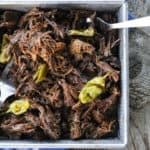 Shredded Crock Pot Mississippi Pot Roast in a square metal pan with whole pepperoncini and meat forks on a blue linen and wooden table.