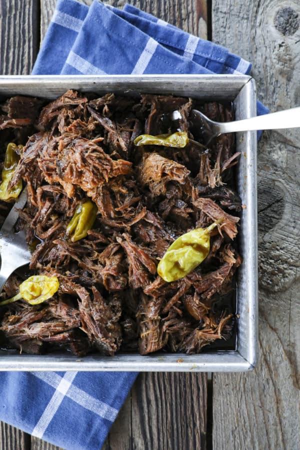 Shredded Crock Pot Mississippi Pot Roast in a square metal pan with whole pepperoncini and meat forks on a blue linen and wooden table. 