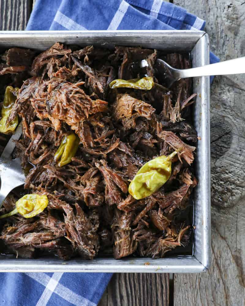 Shredded Crock Pot Mississippi Pot Roast in a square metal pan with whole pepperoncini and meat forks on a blue linen and wooden table.