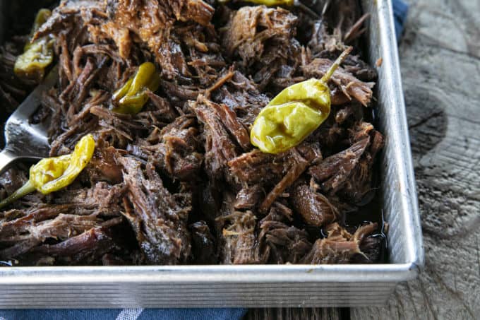 Shredded Crock Pot Mississippi Pot Roast in a square metal pan with whole pepperoncini and meat forks on a blue linen and wooden table.