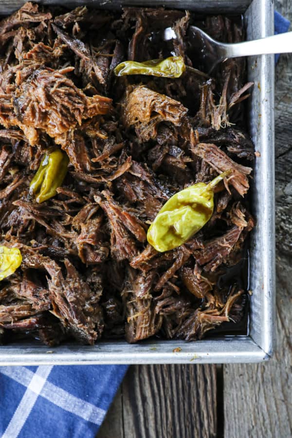 Shredded Crock Pot Mississippi Pot Roast in a square metal pan with whole pepperoncini and meat forks on a blue linen and wooden table.