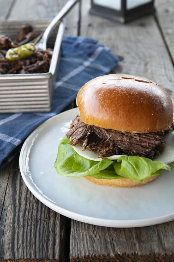 Shredded Crock Pot Mississippi Pot Roast served on a sandwich roll with provolone cheese and butter lettuce on a white plate with a blue linen and wooden table.