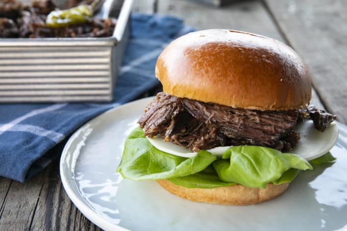 Shredded Crock Pot Mississippi Pot Roast served on a sandwich roll with provolone cheese and butter lettuce on a white plate with a blue linen and wooden table.