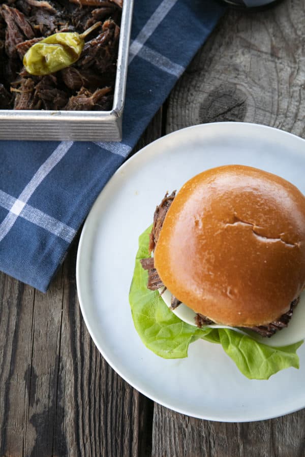 Shredded Crock Pot Mississippi Pot Roast served on a sandwich roll with provolone cheese and butter lettuce on a white plate with a blue linen and wooden table.