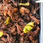 Shredded Crock Pot Mississippi Pot Roast in a square metal pan with whole pepperoncini and meat forks on a blue linen and wooden table.