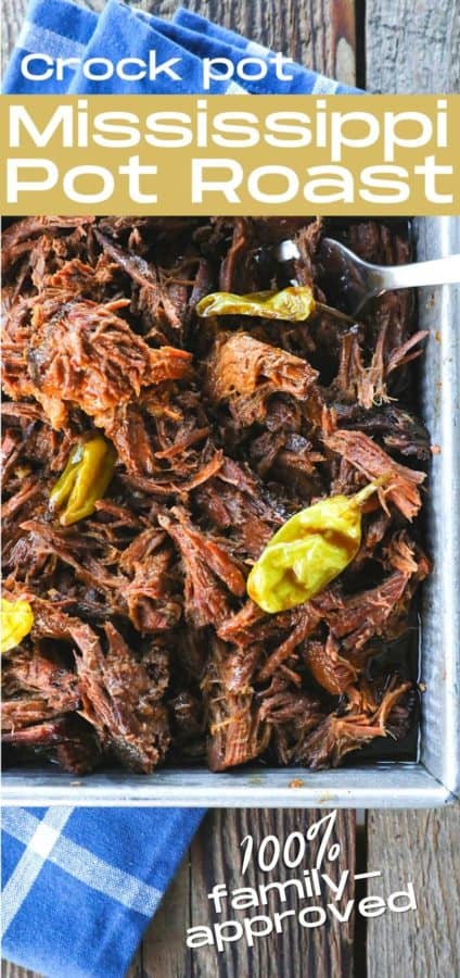 Shredded Crock Pot Mississippi Pot Roast in a square metal pan with whole pepperoncini and meat forks on a blue linen and wooden table.