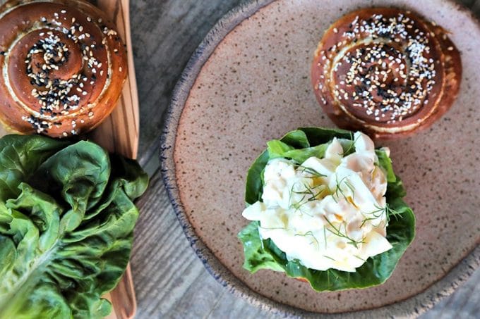 Dill Pickle Egg Salad on lettuce and pretzel roll, brown speckled pottery plate, lettuce leaves, wooden cutting board, rustic wood table, horizontal